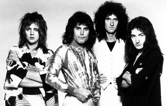 Roger Taylor, Freddie Mercury, Brian May y John Deacon posan de pie en una foto promocional en blanco y negro de Queen en 1975