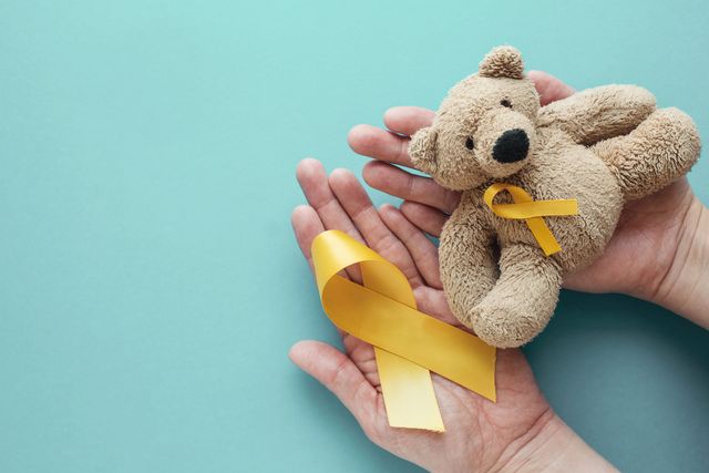 Unas manos sostienen un oso de peluche con el lazo dorado del cáncer infantil