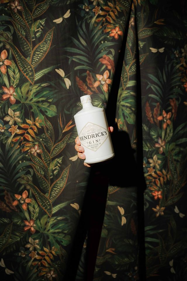 Una mano sostiene una botella de Another Hendrick's frente a una cortina estampada