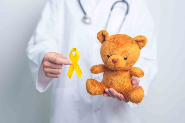 Una mano con bata médica sostiene un lazo dorado junto a un oso de peluche