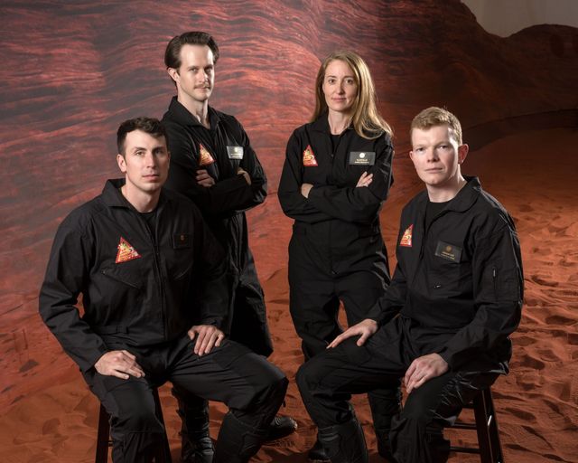 Ross Elder, Matthew Montgomery, Ellen Ellis y James Spicer, con overoles negros y el parche de CHAPEA, posan ante un fondo que imita el paisaje rojizo de Marte