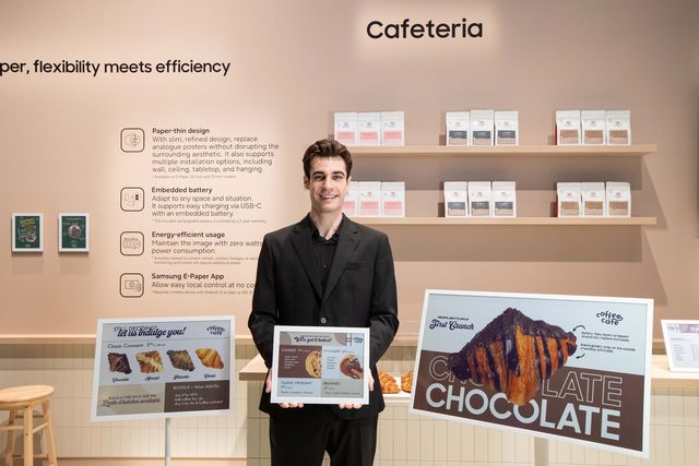 Un hombre sostiene una pantalla Samsung Color E-Paper en una cafetería con varios pósteres digitales