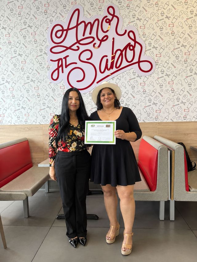 María Gil y Solanyi Cabrera muestran un certificado bajo el letrero «Amor al Sabor» de Pollos Victorina