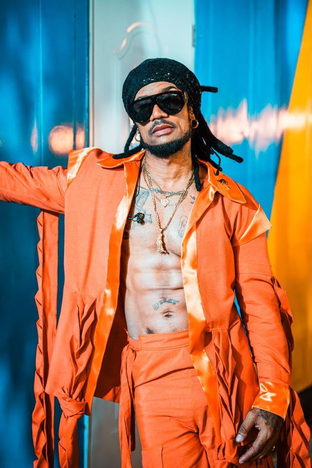 Vakeró posa con un traje naranja de flecos, pañuelo negro y gafas oscuras frente a una pared azul