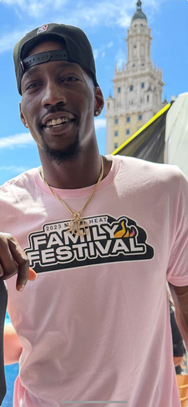 Bam Adebayo sonríe con gorra negra y camiseta rosada del Miami Heat Family Festival, con la Freedom Tower de fondo