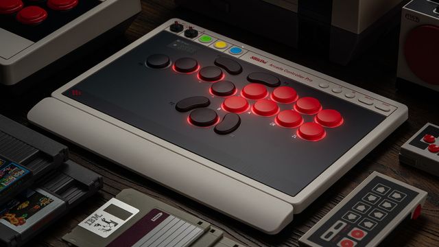 El 8BitDo Arcade Controller Pro, un mando arcade solo de botones con luces rojas, sobre una mesa junto a consolas y mandos retro