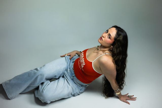 Caluu C. recostada sobre un fondo gris claro, con camiseta roja y jeans