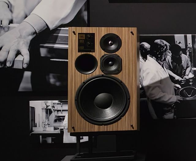 Bocina JBL L100 Classic 80 en madera, edición por los 80 años de la marca, frente a fotos de archivo de su fábrica