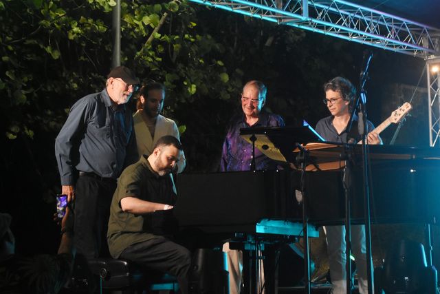 Un pianista toca un piano de cola mientras otros músicos, entre ellos un bajista, lo rodean en la tarima del Festival de Jazz Restauración