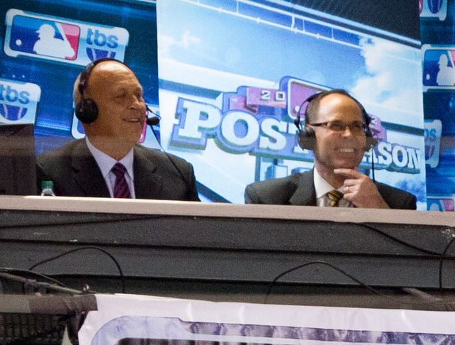 Ernie Johnson, con audífonos y lentes, sonríe en la cabina de transmisión de TBS junto a Cal Ripken Jr. en la postemporada de 2012