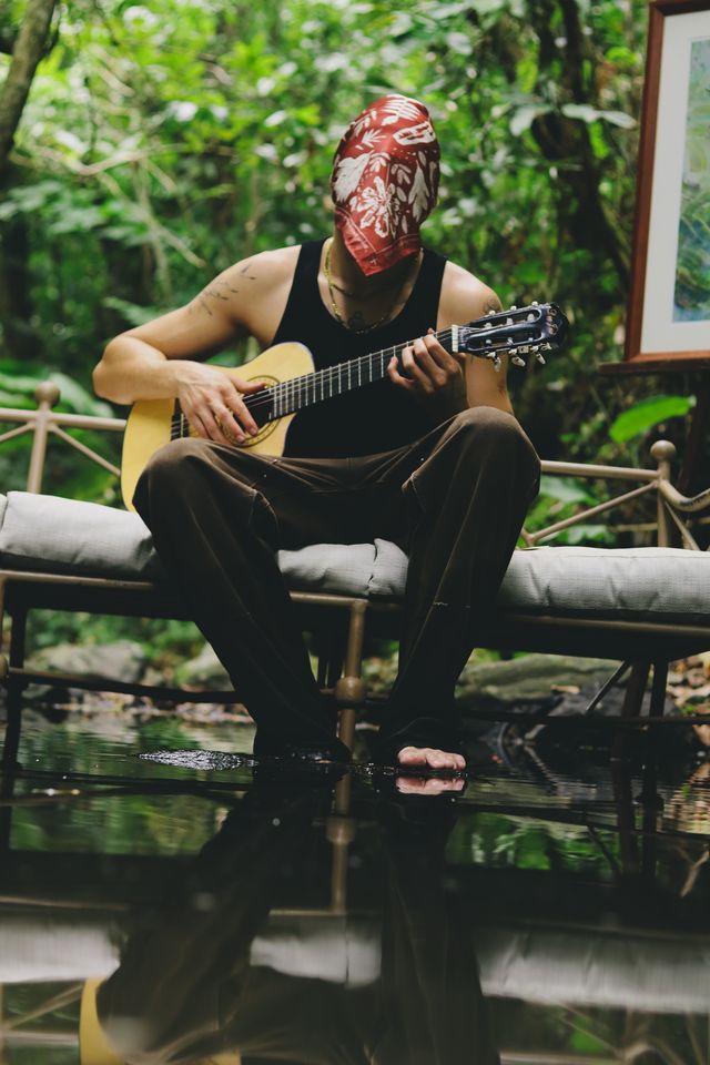 Alzérreca toca la guitarra con el rostro cubierto por un pañuelo rojo, rodeado de vegetación