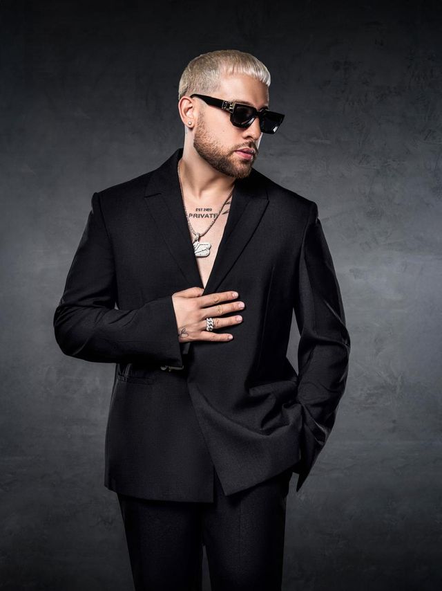 Mark B con traje negro y gafas oscuras, con la mano en el pecho