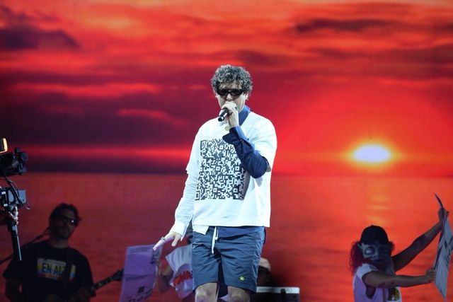Un integrante de Rawayana canta frente a una pantalla con un atardecer en el mar
