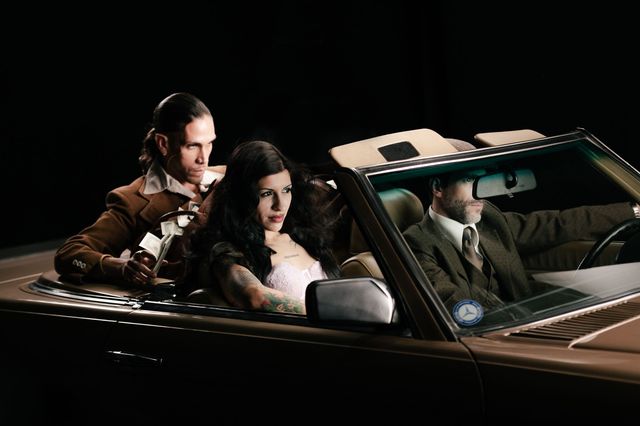 Cazzu en el asiento trasero de un convertible antiguo junto a dos hombres trajeados
