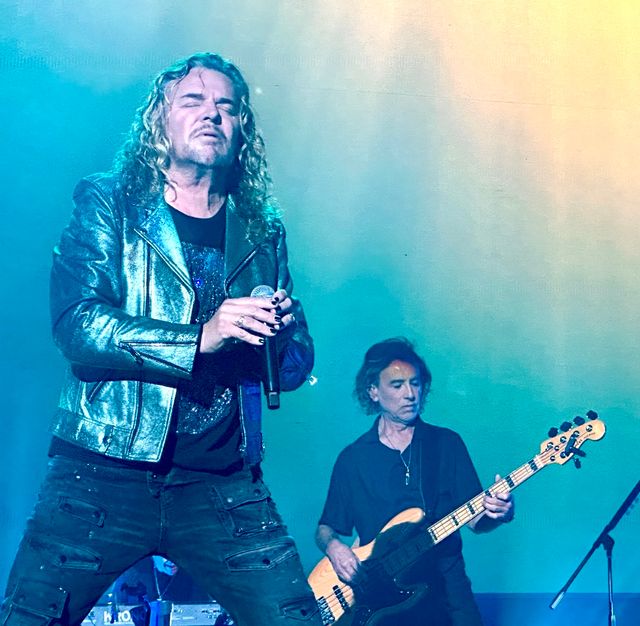 Fher Olvera canta con chaqueta de cuero plateada mientras Juan Calleros toca el bajo en un concierto de Maná en Denver en 2023