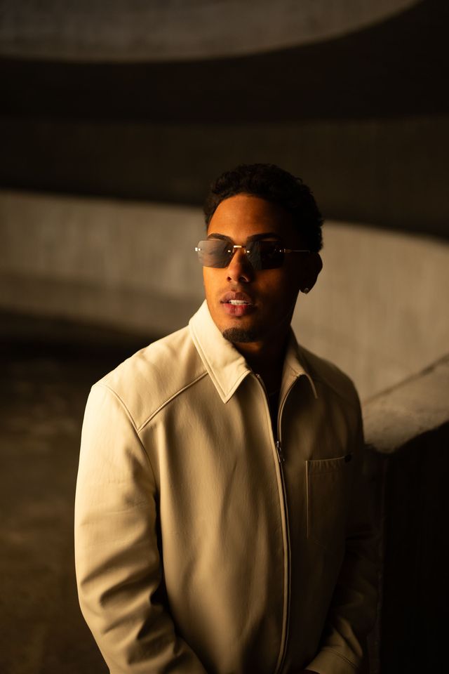 Myke Towers con gafas oscuras y chaqueta clara en un pasillo iluminado