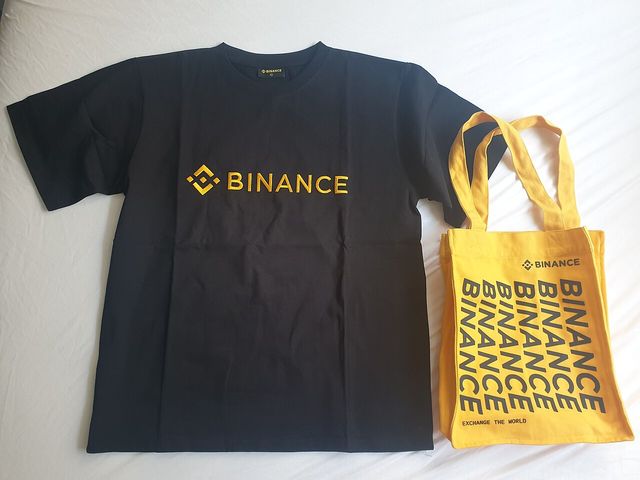 Una camiseta negra y una bolsa amarilla con el logo de Binance sobre una sábana blanca