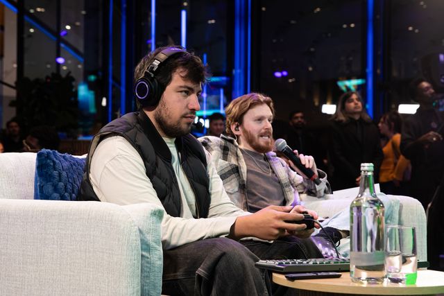 Dos jugadores con control y audífonos en un sofá durante una partida, uno de ellos con micrófono