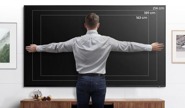 Un hombre con los brazos abiertos frente a un televisor de gran formato con medidas de pantalla marcadas