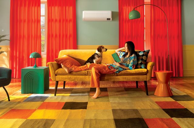 Una mujer lee en un sofá amarillo junto a su perro, con un aire acondicionado Samsung en la pared