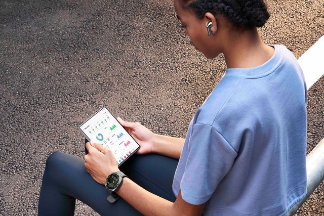 Una joven sentada al aire libre revisa datos de salud en un teléfono plegable Galaxy con su reloj y audífonos