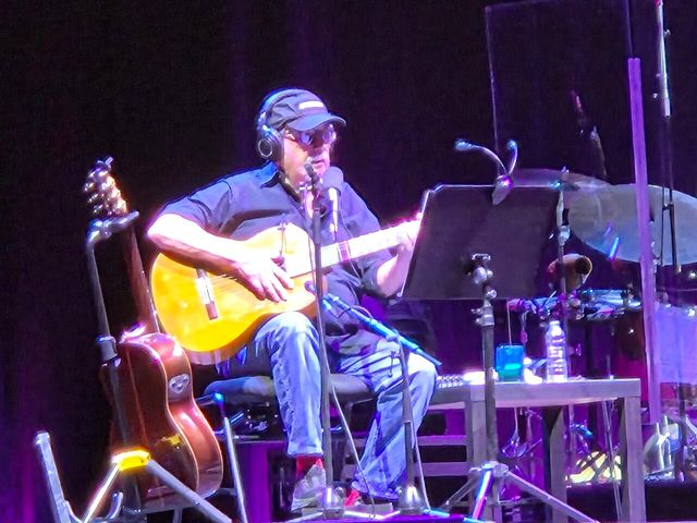 Silvio Rodríguez, con gorra y audífonos, toca la guitarra sentado en el escenario