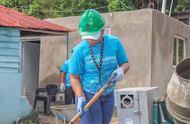 Una voluntaria con casco verde trabaja con una pala junto a una mezcladora de concreto