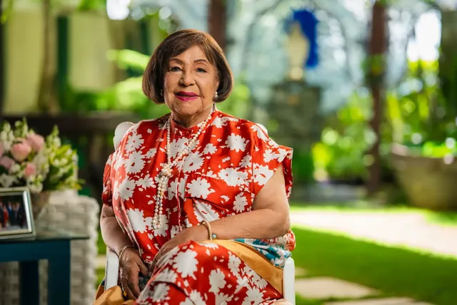 Camelia Ureña, con vestido rojo de flores blancas, sentada en un jardín